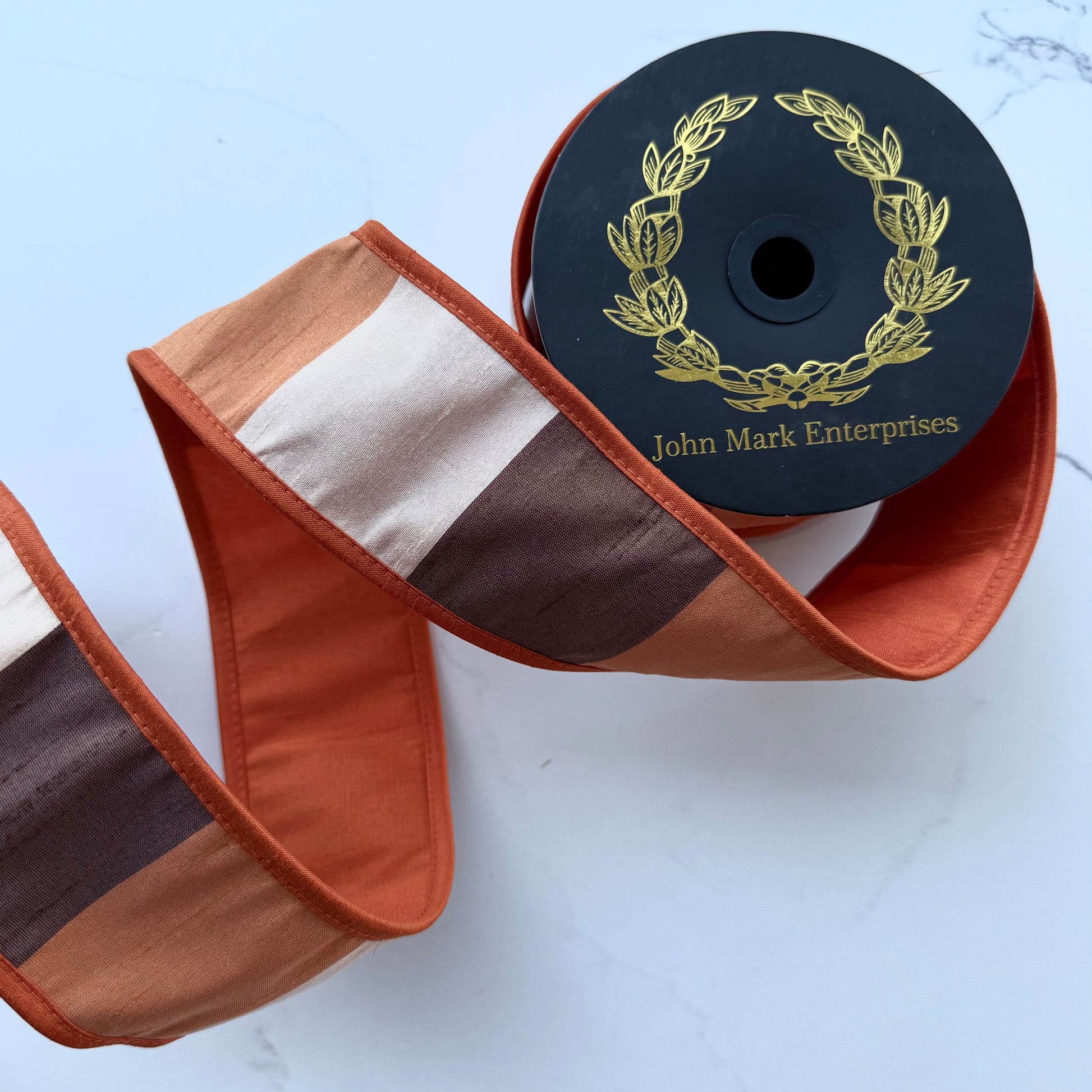 roll of ribbon with alternating stripes of rust orange, cream, and dark brown, slightly unrolled to show solid orange backing on a light surface. The spool is black with a gold laurel wreath logo and the brand name 'John Mark Enterprises'.