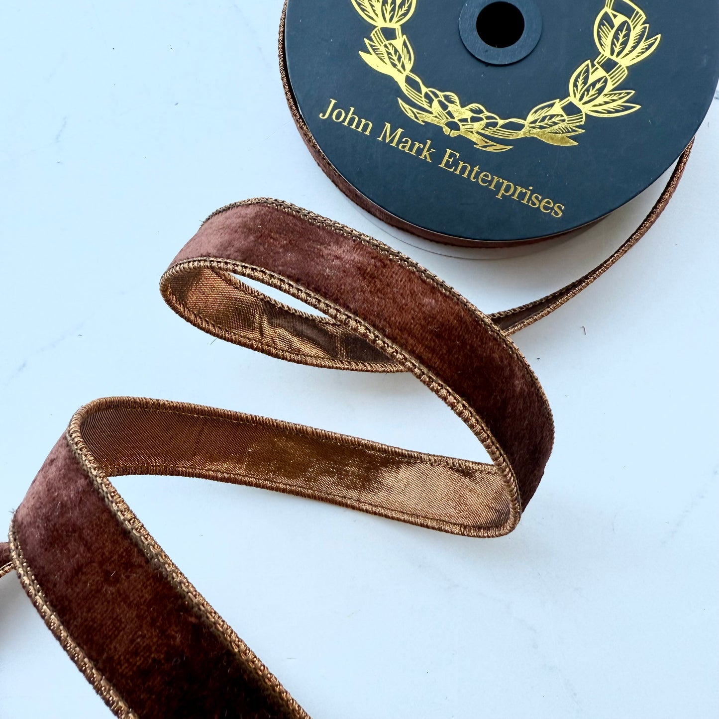 A close-up of a spool of brown velvet ribbon with with coppery colored backing and the brand name 'John Mark Enterprises' printed on the spool. The ribbon is draped in a loose spiral to show both sides