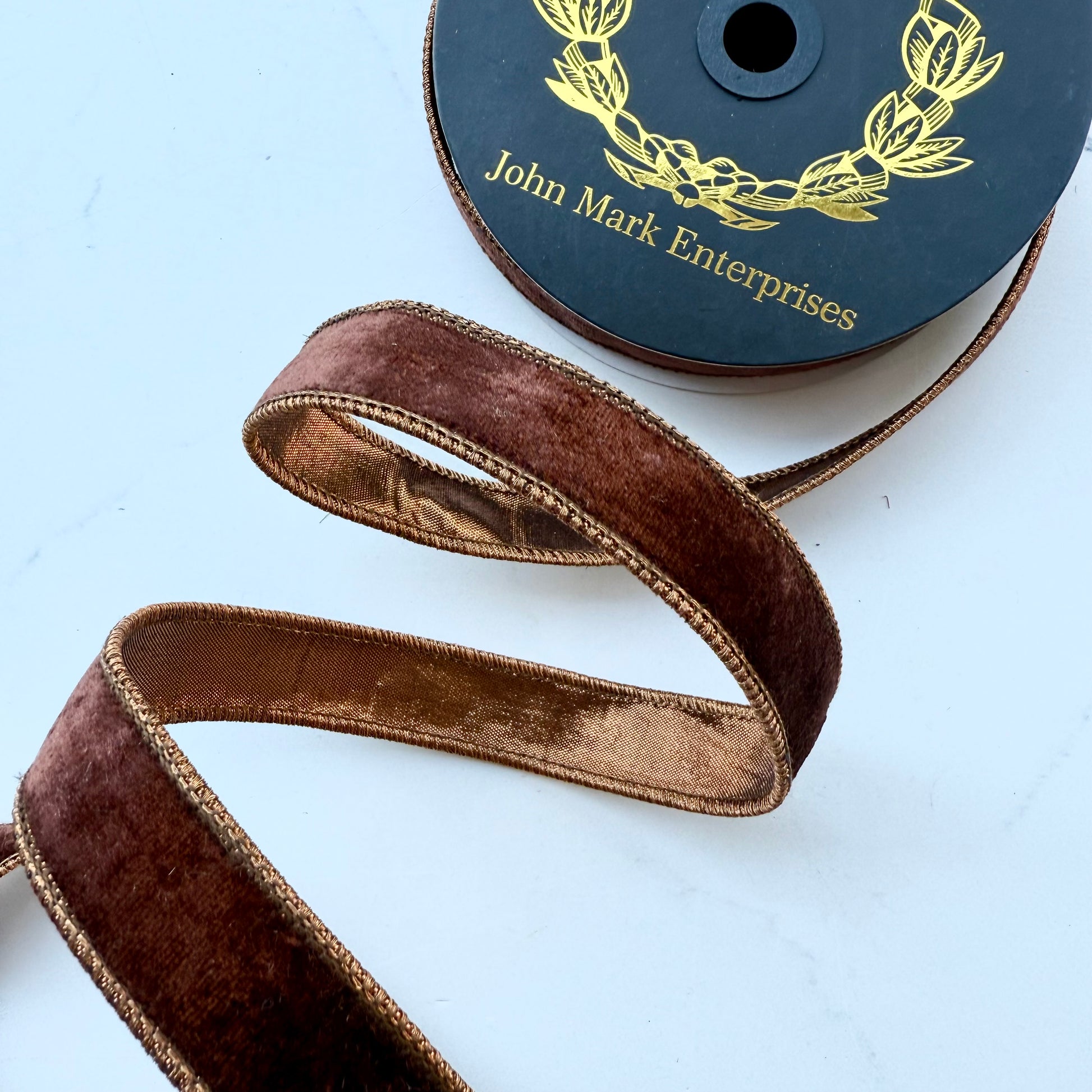 A close-up of a spool of brown velvet ribbon with with coppery colored backing and the brand name 'John Mark Enterprises' printed on the spool. The ribbon is draped in a loose spiral to show both sides