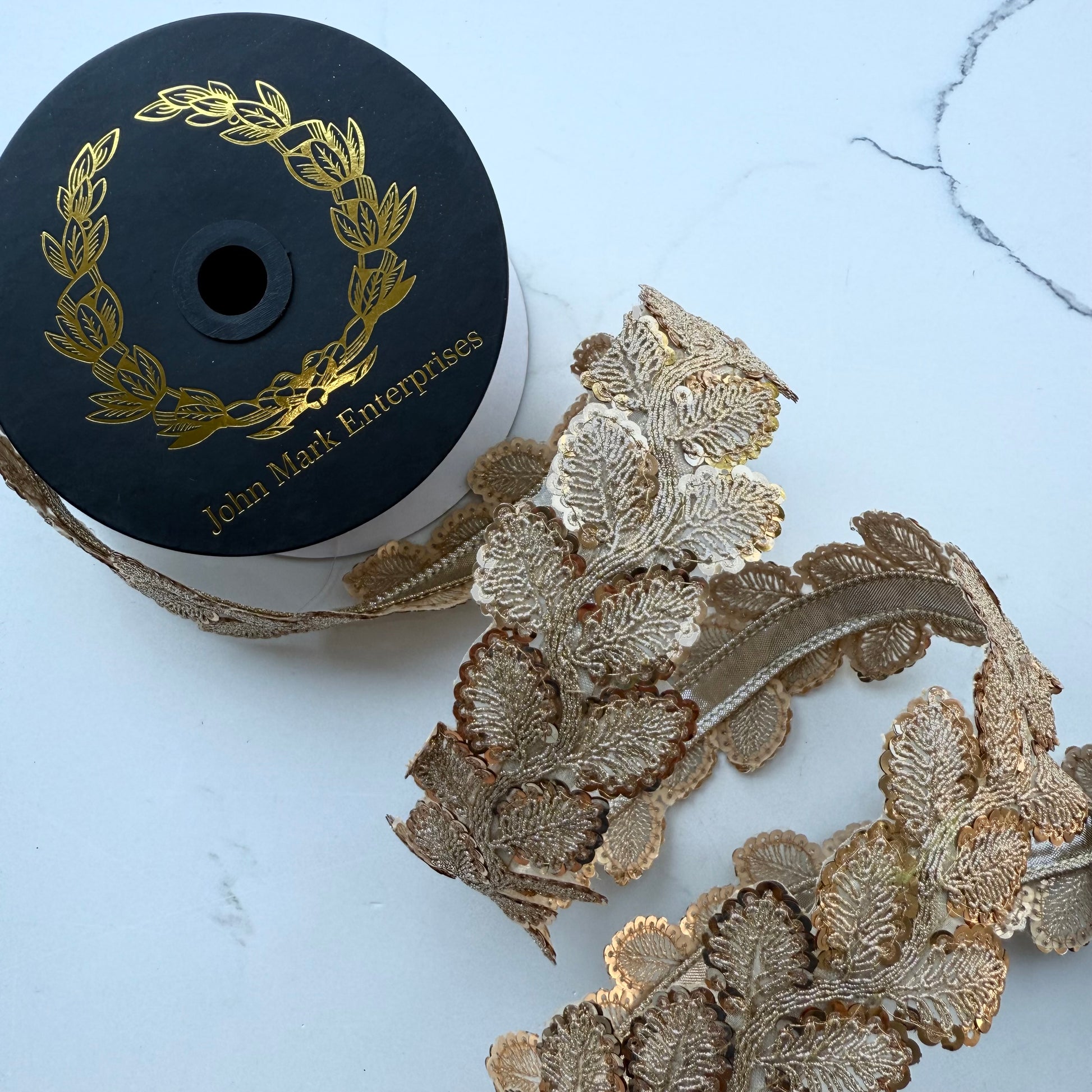 Gold and beige sequin leaf ribbon on a spool with a black label reading 'John Mark Enterprises', shown on a light marble-like surface.