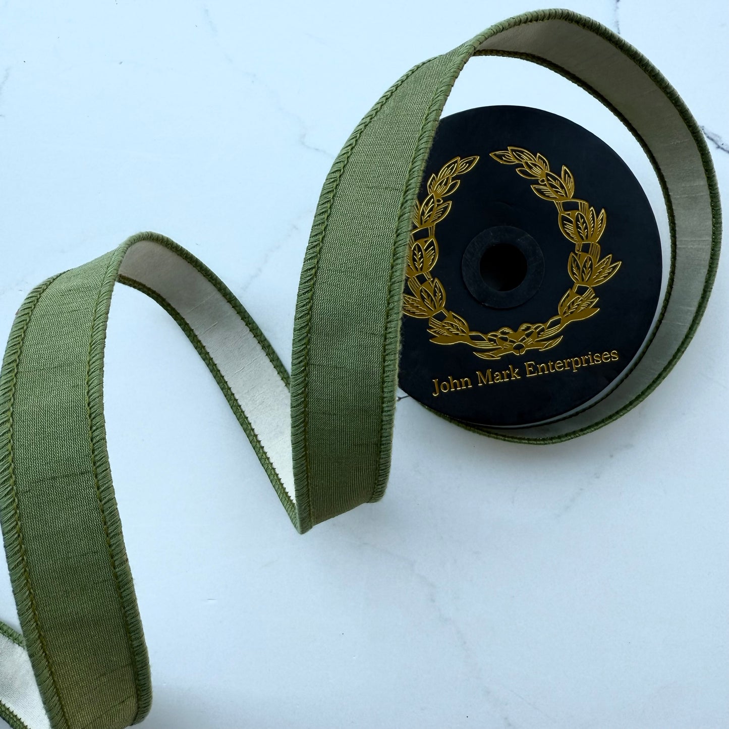 Olive green ribbon with a ivory backing, coiled on a black spool with gold lettering that reads 'John Mark Enterprises'.