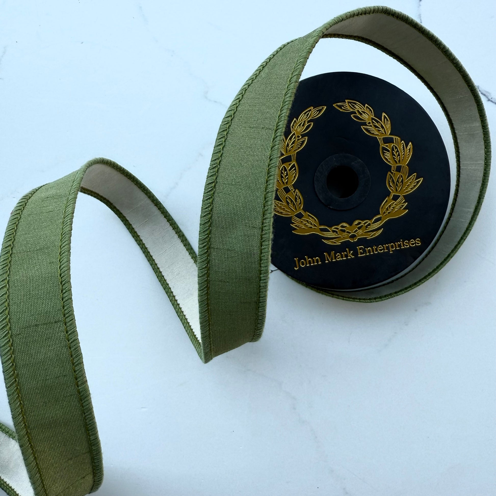 Olive green ribbon with a ivory backing, coiled on a black spool with gold lettering that reads 'John Mark Enterprises'.