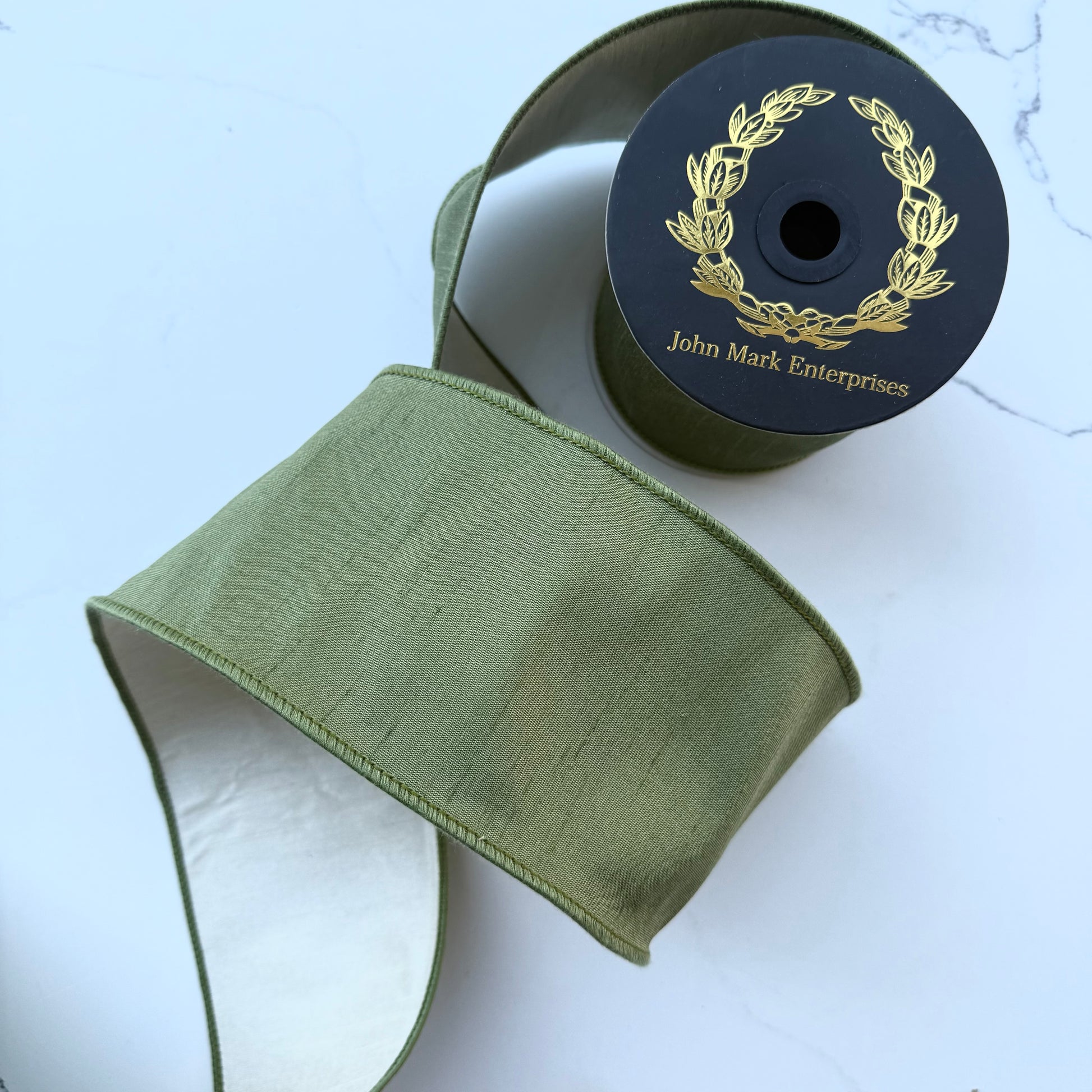 Olive green ribbon slightly unrolled to show ivory backing on a black spool with label printed with a gold laurel wreath and the brand name 'John Mark Enterprises'