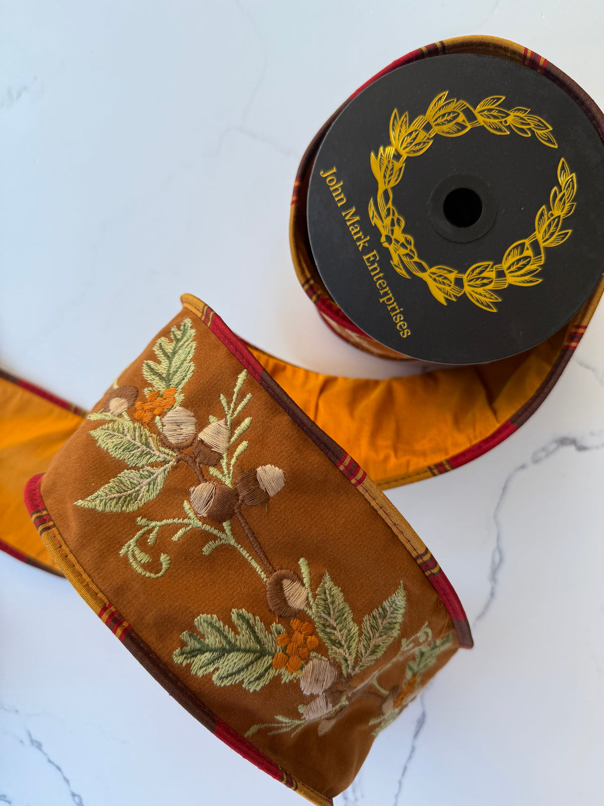 An ornate embroidered ribbon spool  features a warm brown base with detailed embroidery of leaves, acorns, and small orange flowers, finished with a red and gold plaid border.