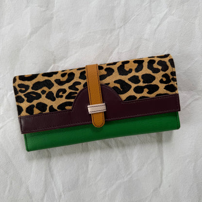 A long wallet with a leopard print top flap, a dark burgundy leather band, a tan strap closure, and a green body, displayed on a wrinkled white fabric background.