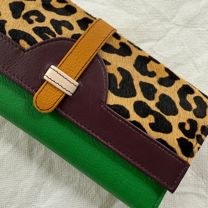 A close-up of a wallet featuring a leopard print flap, a tan leather strap, and a bright green body, laid on a light textured fabric background.