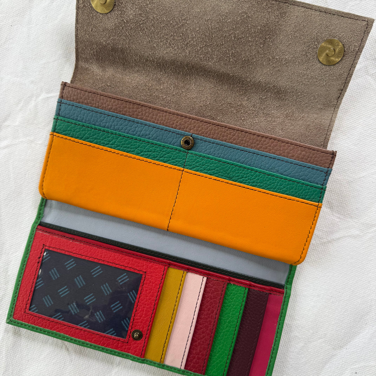 An open leather wallet  made from multiple bright colors, including orange, green, red, pink, yellow, and blue, laid flat on a white textured fabric background.