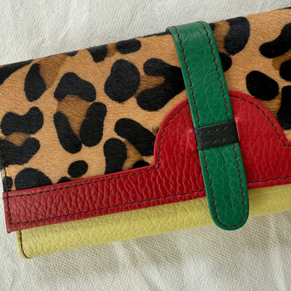 A close-up of a multicolor leather clutch wallet featuring a leopard print pattern, a red leather band, a green strap, and a pale yellow base