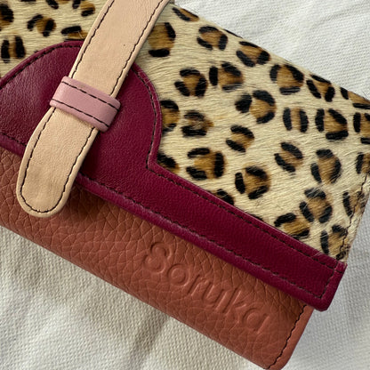 A close-up of a Soruka wallet featuring a leopard print hair-on-hide panel, a magenta leather trim, and mauve body