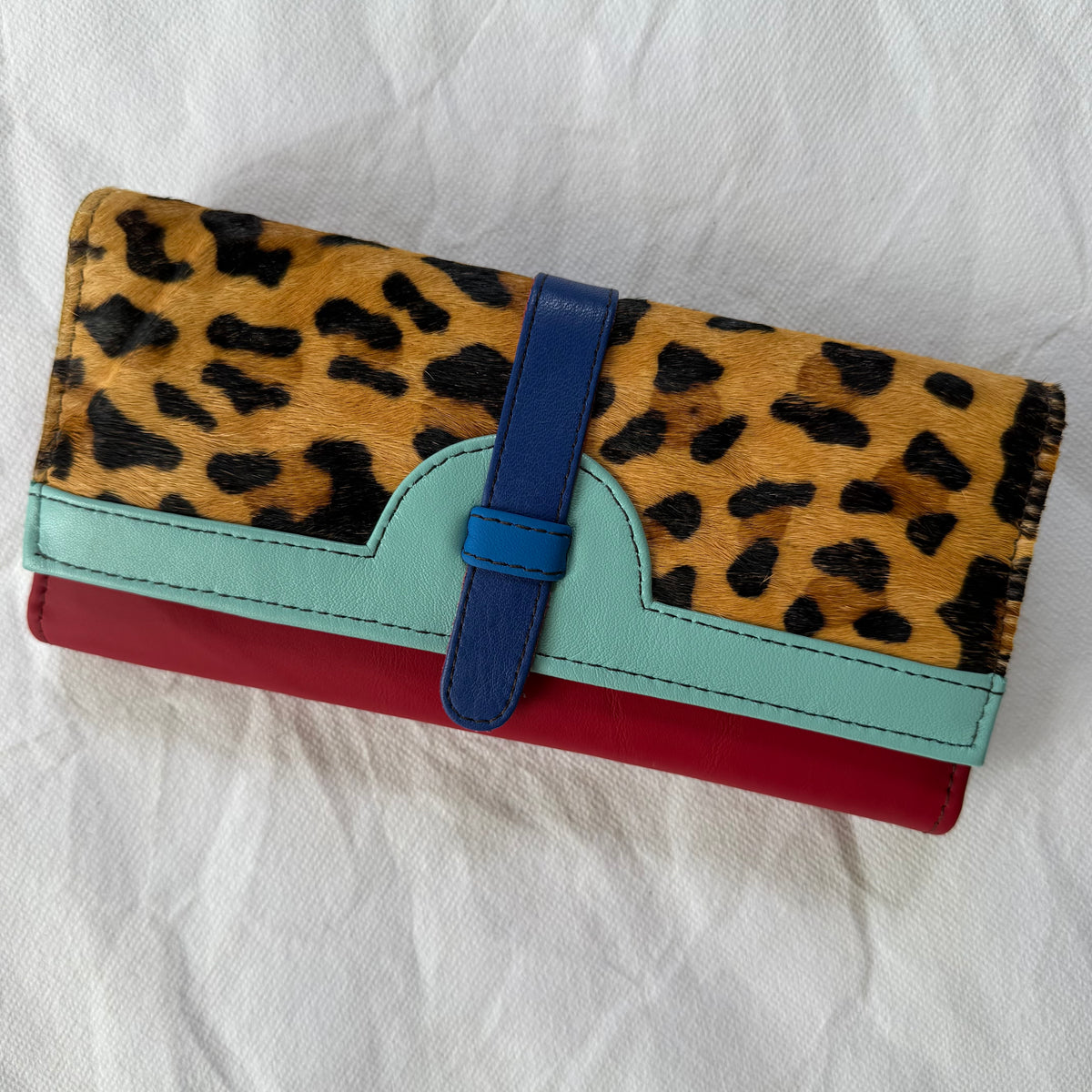 A multi-colored wallet featuring a leopard print top flap, a light blue leather trim, a blue strap closure, and a red base, displayed on a white fabric background.