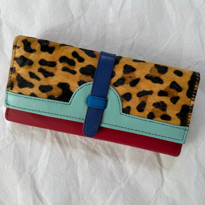 A multi-colored wallet featuring a leopard print top flap, a light blue leather trim, a blue strap closure, and a red base, displayed on a white fabric background.