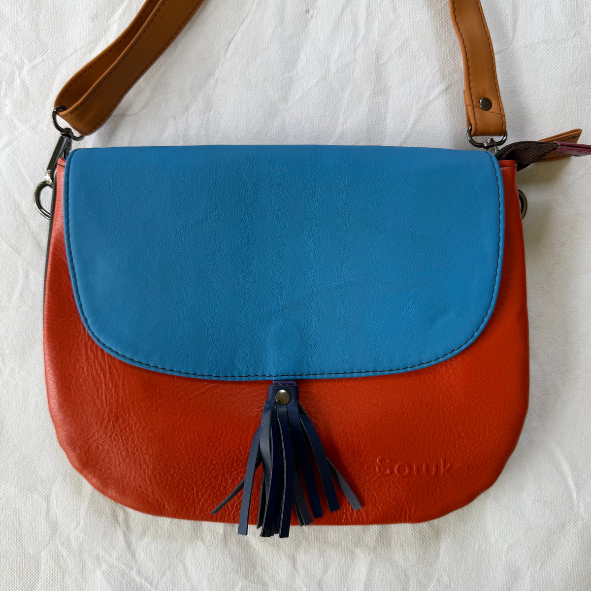 A color-blocked leather shoulder bag with a bright blue top flap, a rust-orange lower body, and dark navy fringe tassels. 