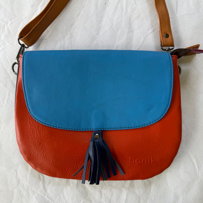 A color-blocked leather shoulder bag with a bright blue top flap, a rust-orange lower body, and dark navy fringe tassels. 