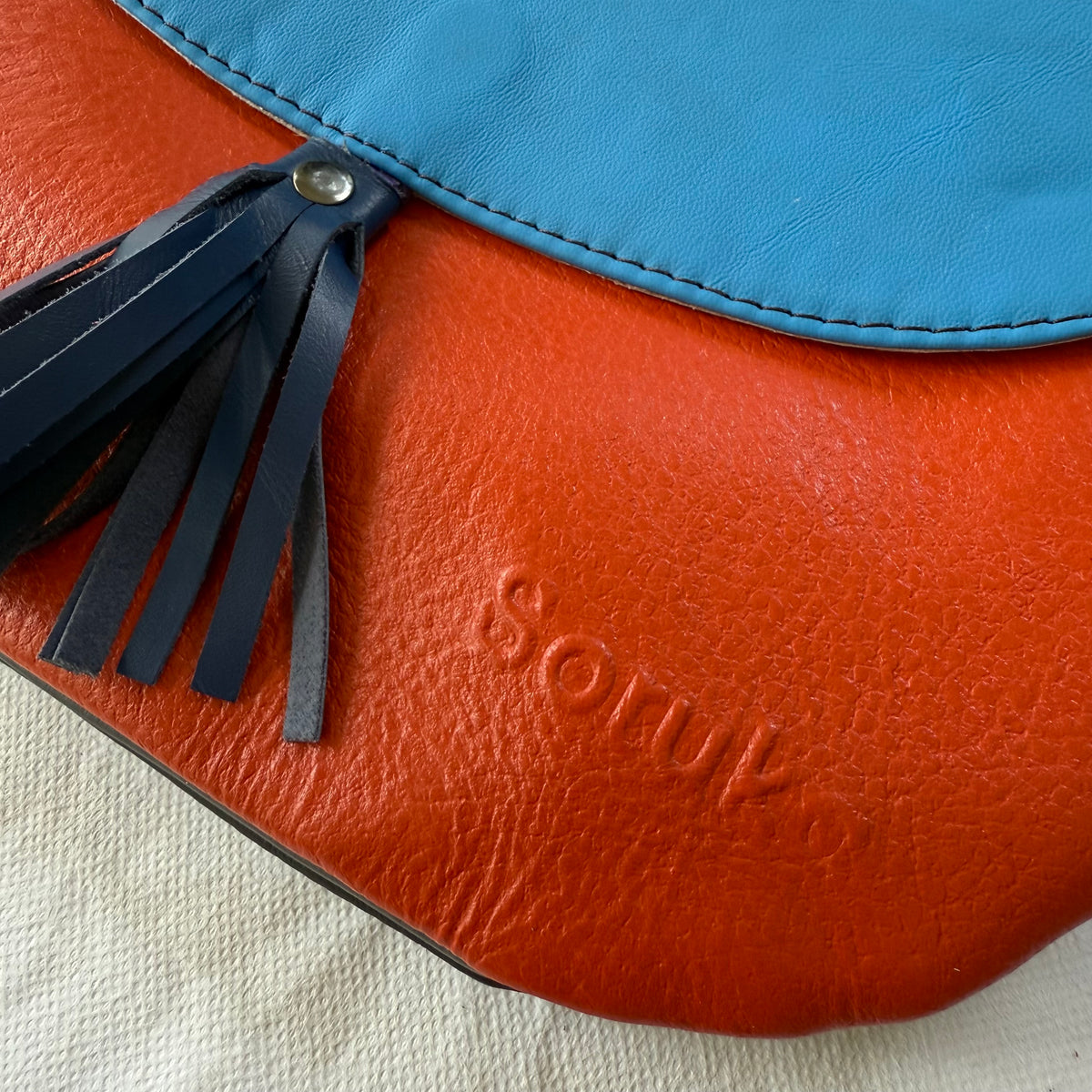 Close-up of a leather bag with a bright orange-red body, a turquoise blue flap, and a dark navy tassel detail. The brand name 'soruka' is embossed on the orange-red leather.