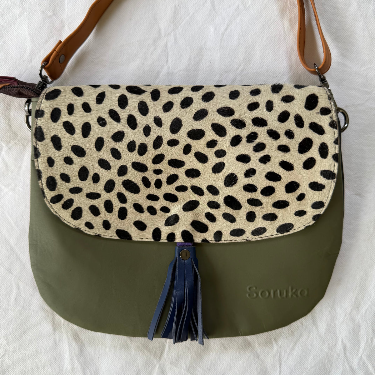 A shoulder bag with a cream and black spotted flap and an olive green body, featuring a navy blue tassel and the name 'Soruka' printed on the front