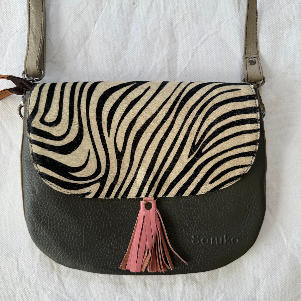 A shoulder bag with a warm grey pebbled leather body, a flap featuring a black and beige zebra print, and a pink tassel accent