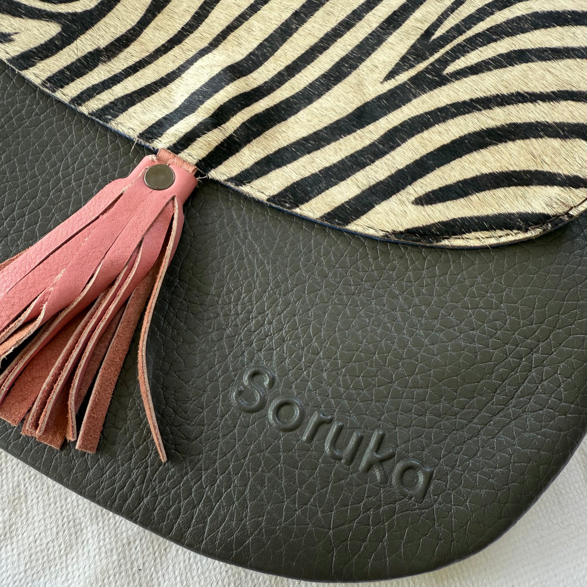 Close-up of a Soruka bag featuring a grey pebbled leather body, a pink leather tassel, and a beige and black zebra-print flap.