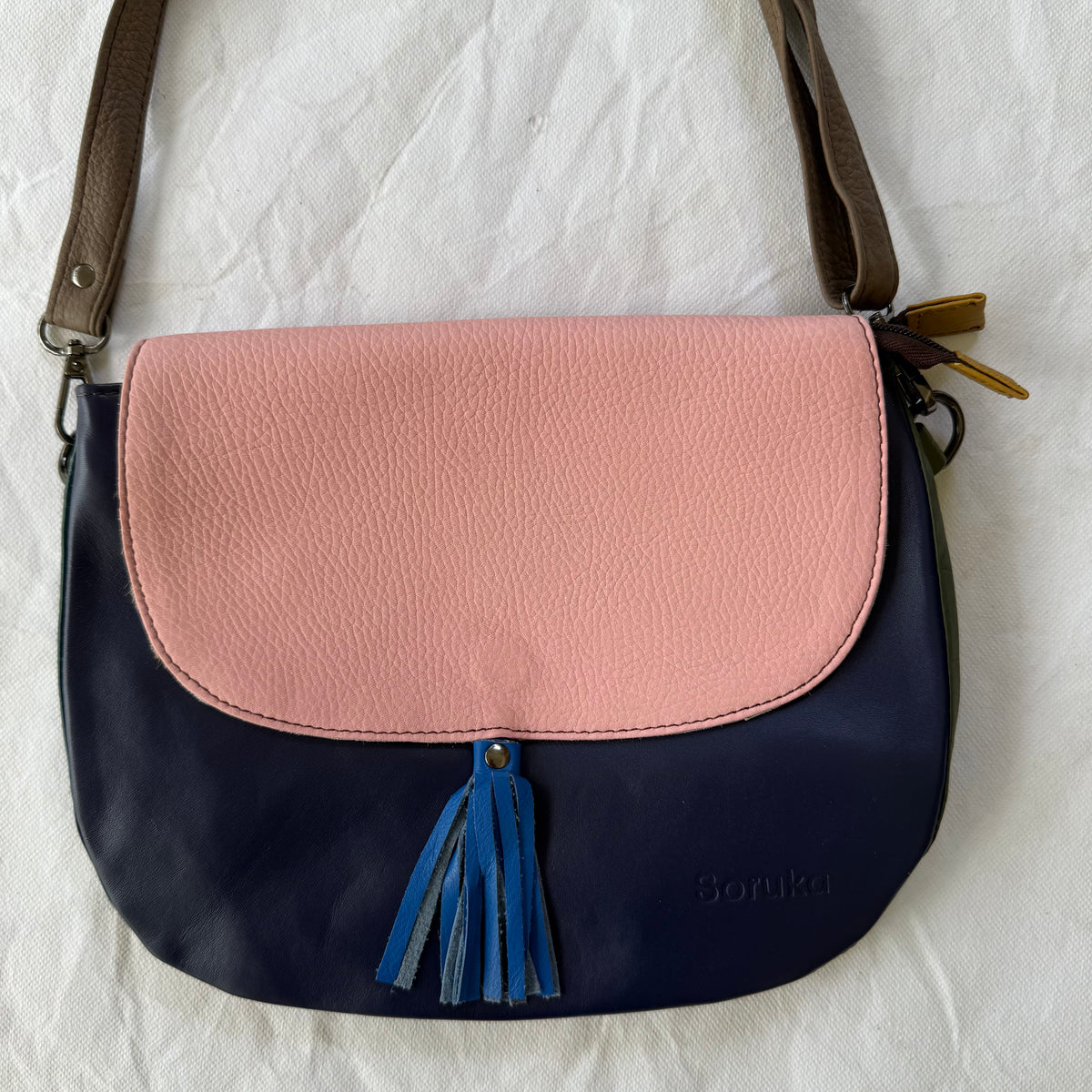 A color-blocked leather shoulder bag with a dusty pink top flap, a navy blue lower body, a blue tassel, and a brown strap, laid flat on a white fabric background.
