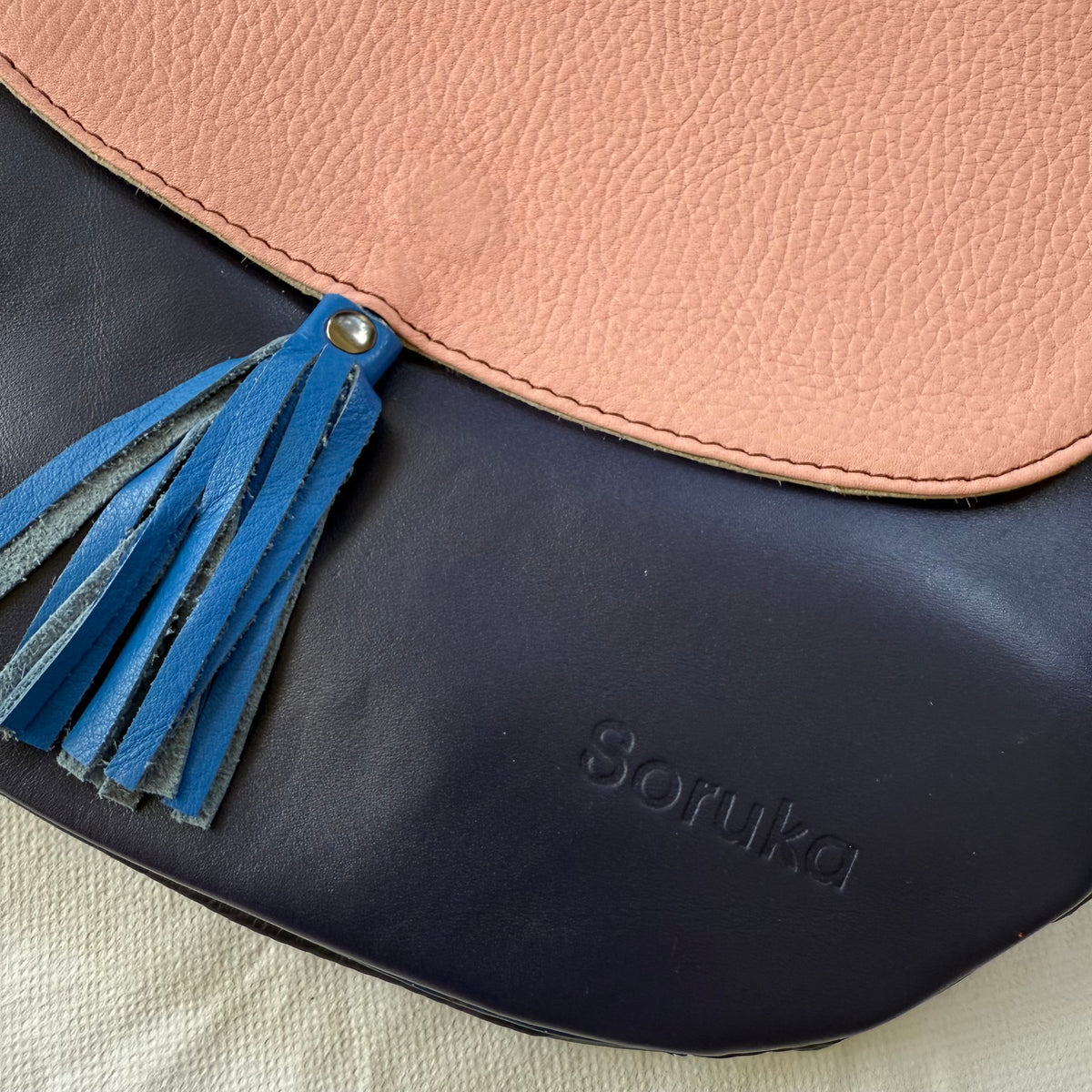 Close-up of a Soruka leather bag with a pale pink-colored top flap and a dark navy blue body, featuring a blue leather tassel detail.