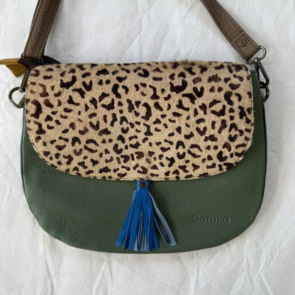A shoulder bag with a green leather body, a faux leopard-print fur flap, and a blue tassel, shown on a white fabric background.