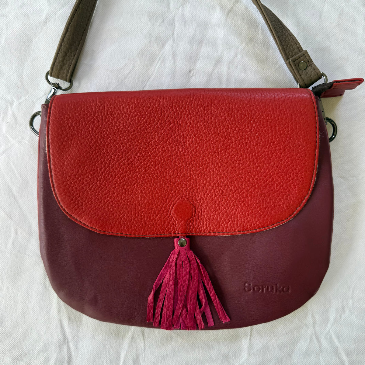 A color-blocked leather shoulder bag with a textured red top flap and a deep burgundy bottom body, featuring a magenta tassel 
