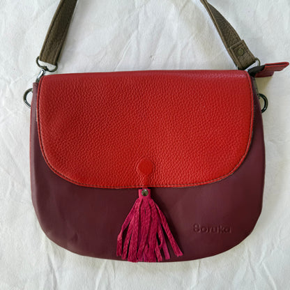 A color-blocked leather shoulder bag with a textured red top flap and a deep burgundy bottom body, featuring a magenta tassel 