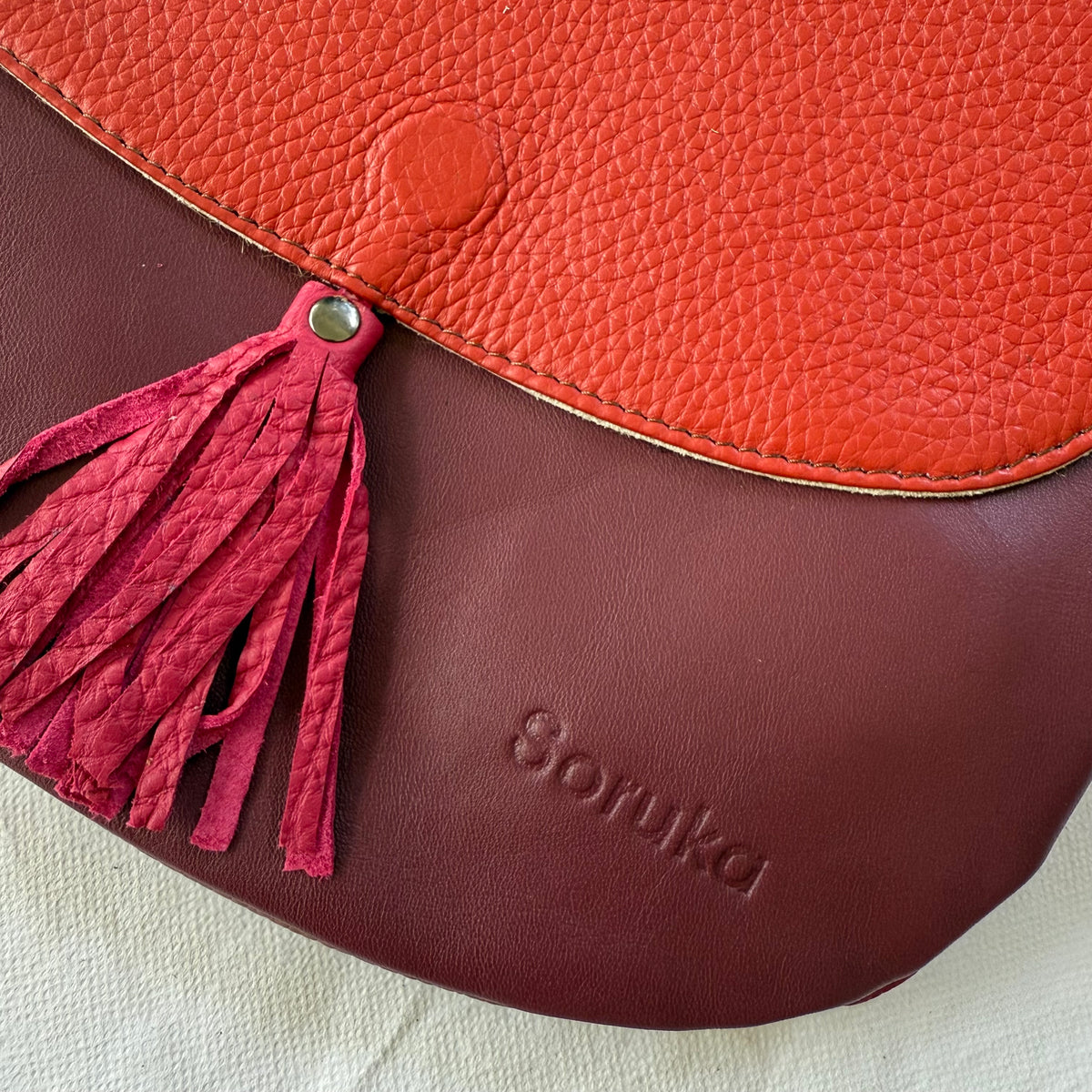 Close-up of a leather shoulder bag with a textured orange-red flap, a dark burgundy body, and a matching fuchsia tassel. The brand name 'Soruka' is embossed on the bag.