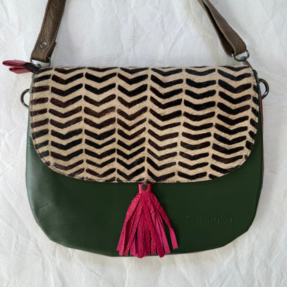 A shoulder bag with a dark green leather body, a brown and beige chevron-patterned flap, and a bright pink tassel, laid flat on a white fabric background.