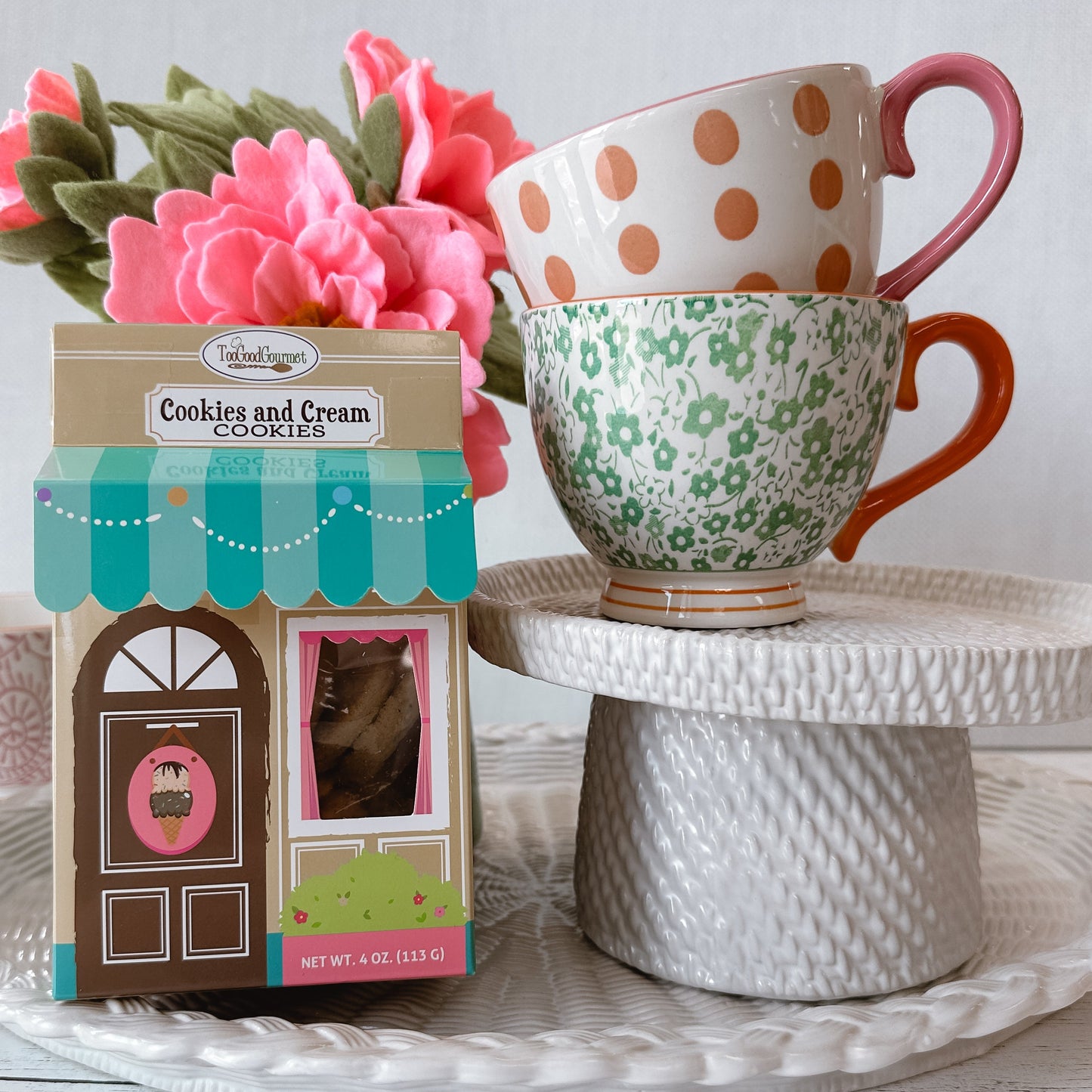 A boxed box of 'Cookies and Cream' cookies sits beside two stacked ceramic mugs on a white pedestal. The box is illustrated like a bakery storefront with a teal awning and pink cupcake graphic.