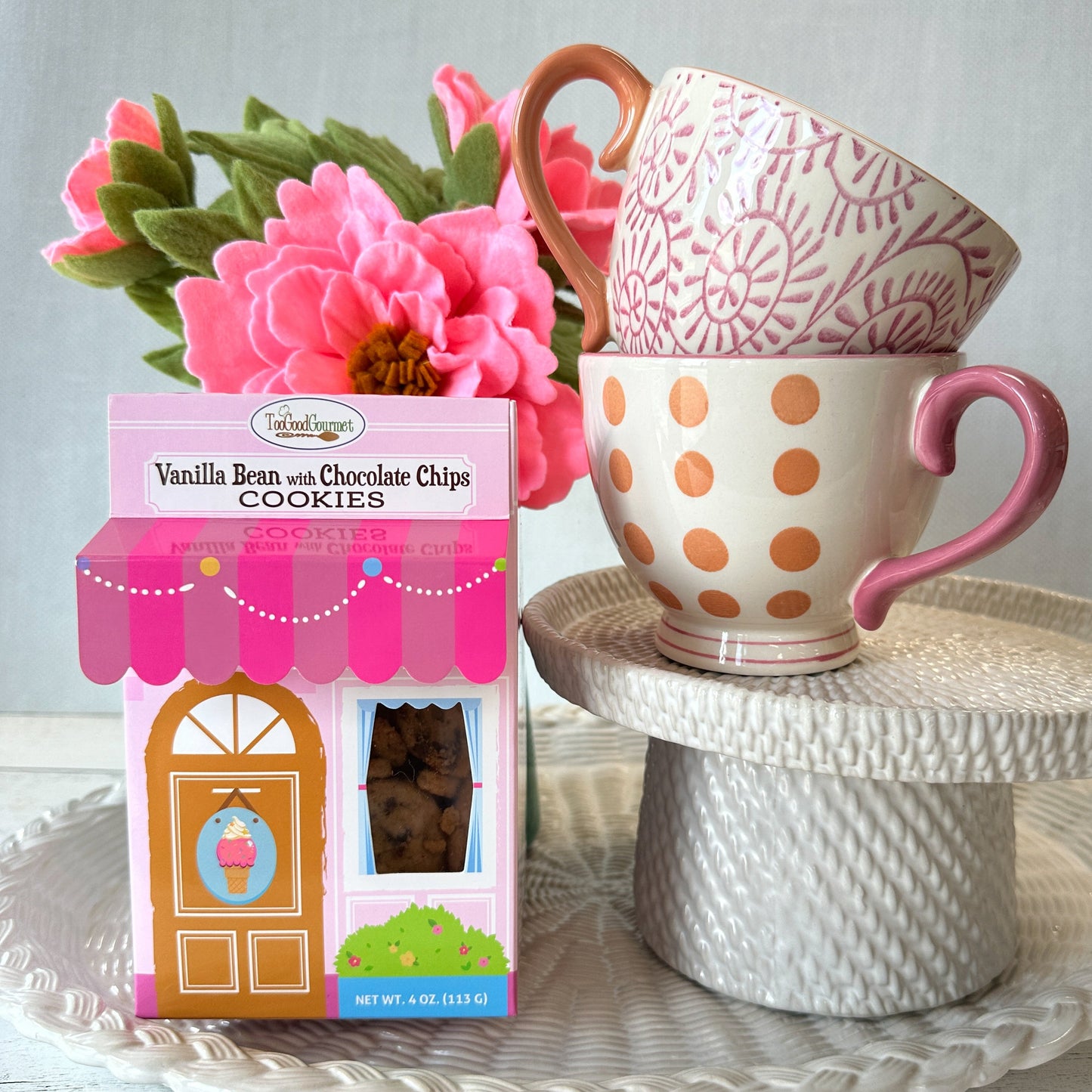 A box of Vanilla Bean and Chocolate Chips Cookies sits next to two stacked ceramic mugs on a white pedestal. The box is pink with a ice cream shop storefront illustration, and the mugs are cream with pink and orange patterns.