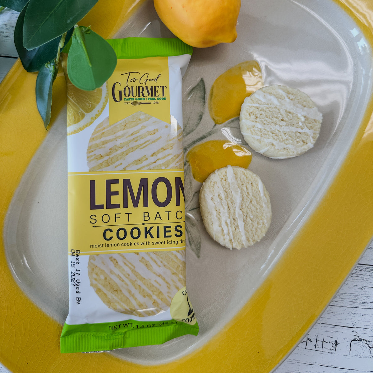 A package of Too Good Gourmet Lemon Soft Batch Cookies placed on a yellow plate with lemon slices, a lemon half, and green leaves, on a white wood surface.