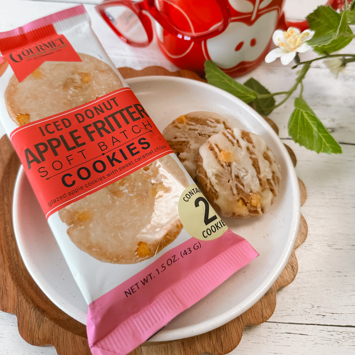 A package of Gourmet Apple Fritter Soft Batch Cookies is shown on a white plate next to two cookies, with a red mug and green leaves in the background.