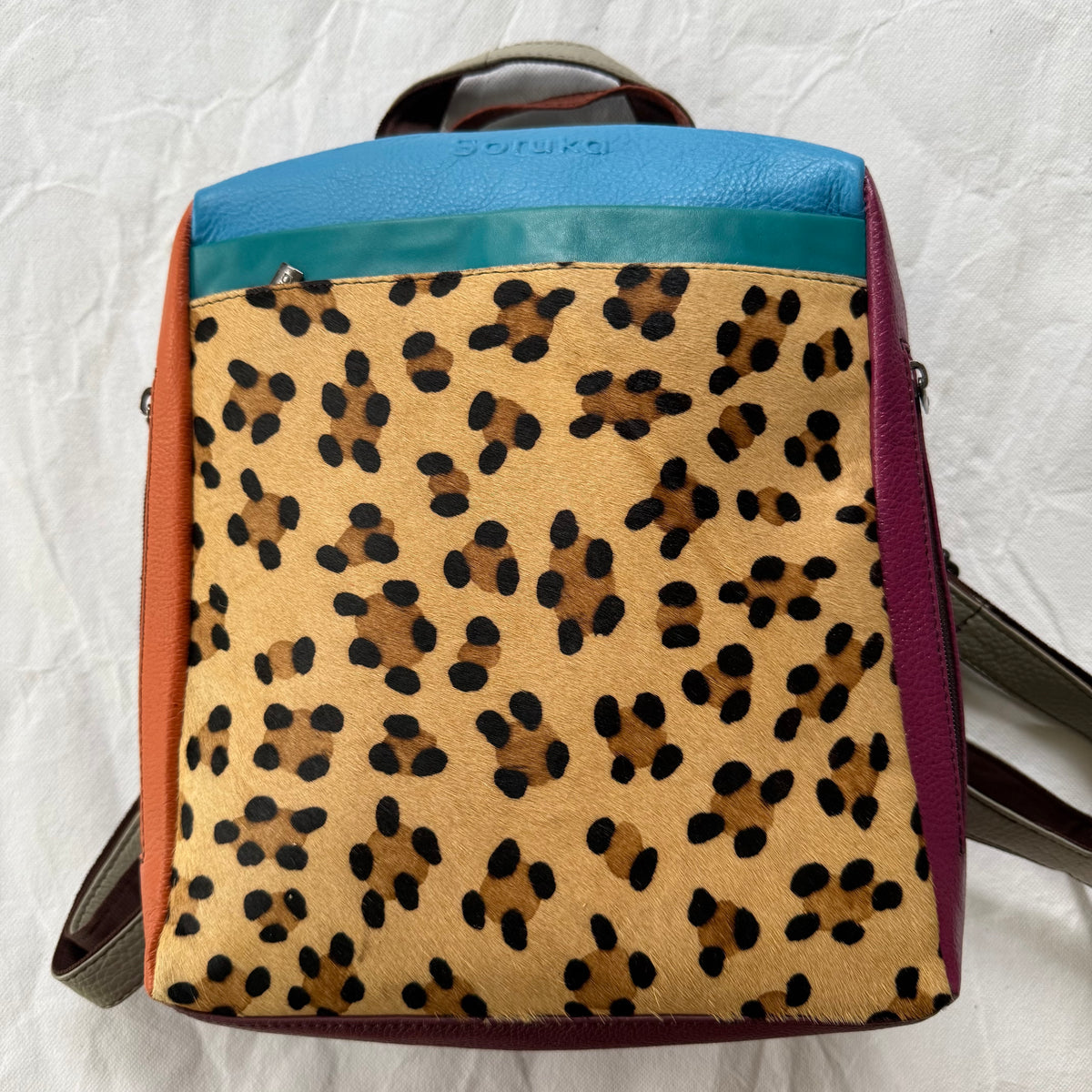 A color-blocked leather backpack featuring a tan and black leopard print front panel, a bright blue top panel, and side panels in orange, magenta, and dark green.