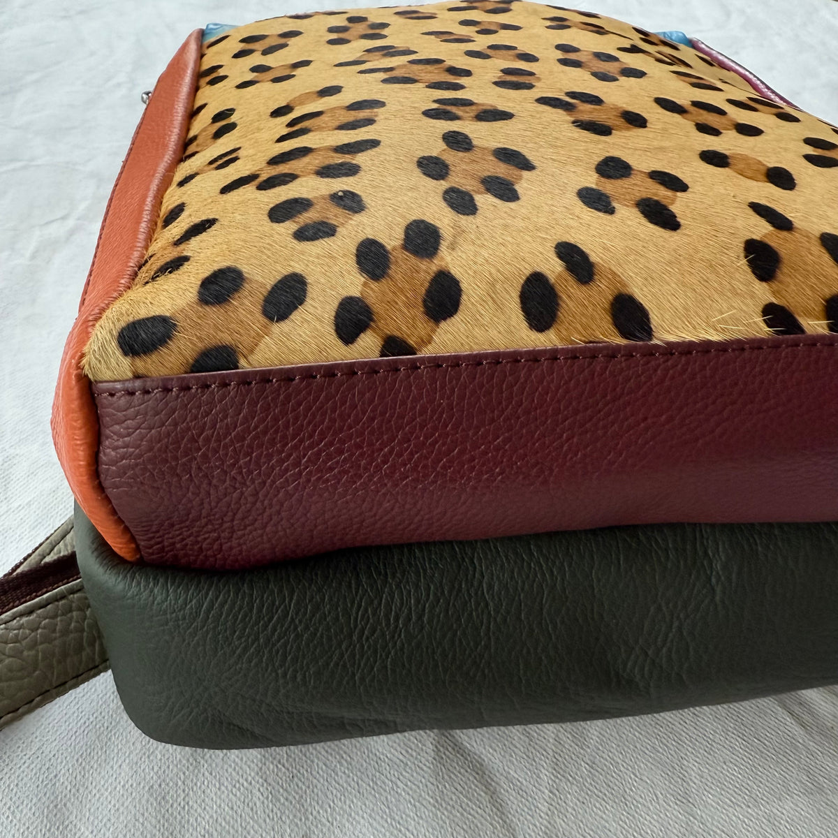A close-up of a handbag showing its layered leather construction, featuring a tan cheetah print top panel with black spots, a reddish-brown leather side panel, and a dark olive green bottom panel.