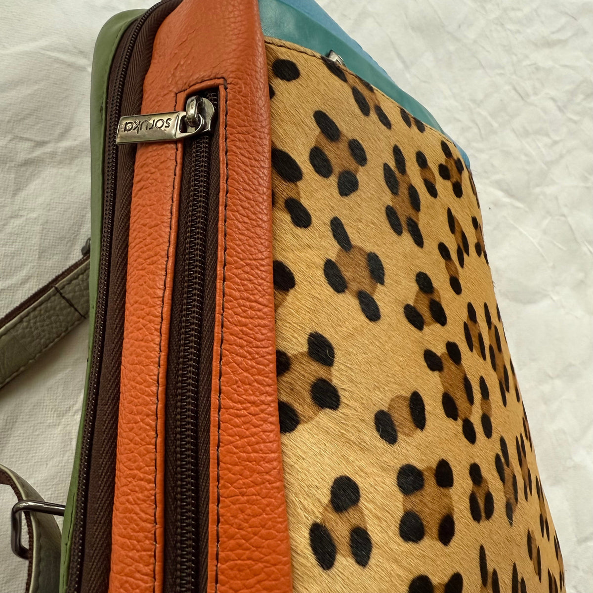 Close-up of a designer handbag showing its side profile, featuring a leopard print hair-on-hide panel, a textured orange leather zipper section, a dark brown zipper, and a green leather strap on a white fabric background.