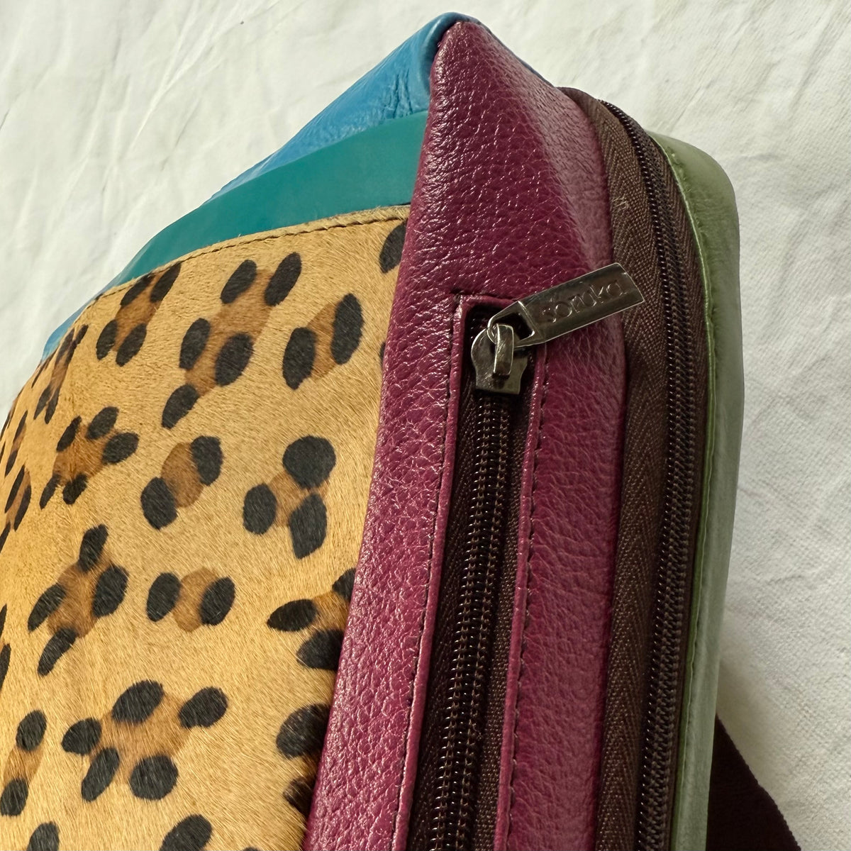 Close-up of a multicolor bag side showing a leopard print panel, magenta pebbled leather, a dark brown zipper, and a teal fabric section on a white textured background.