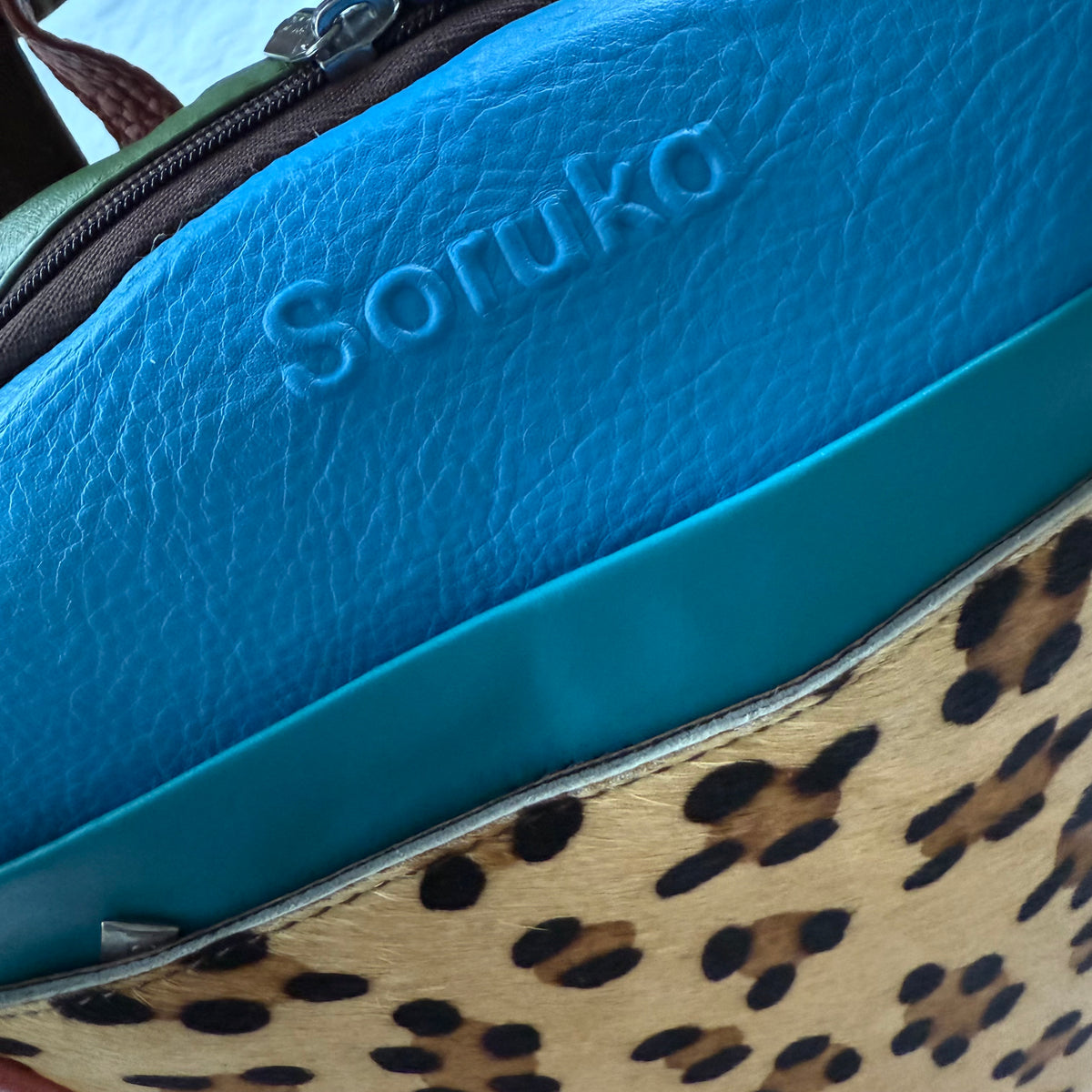Close-up of top view of a colorful bag showing a bright blue textured leather section with the embossed brand name 'Soruka', a teal trim band, and a tan-and-black leopard print panel.