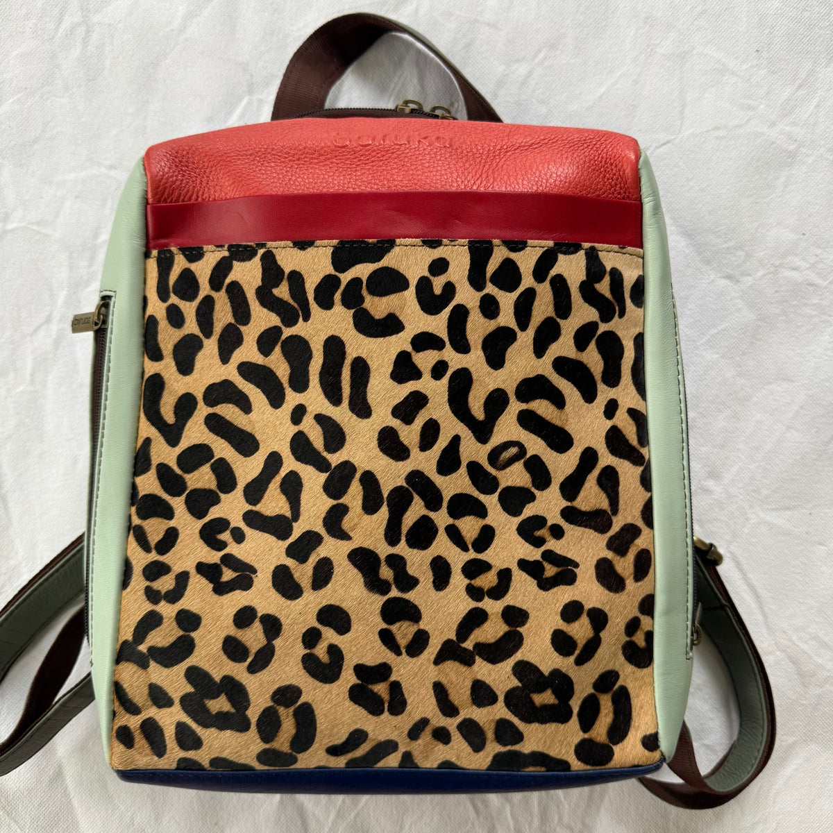 A square leather backpack with a leopard print pattern on the front, a coral red top panel, pale mint green side panels, and dark brown straps