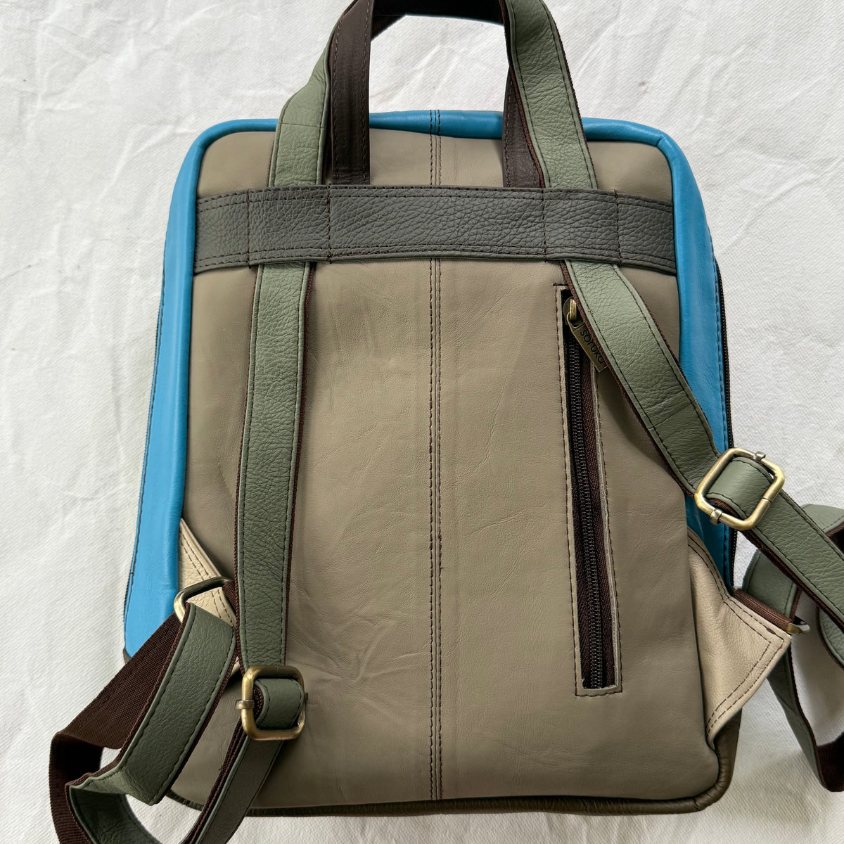 The back view of a leather backpack in taupe and olive green with bright blue side panels, featuring dual top handles, adjustable shoulder straps, and a vertical zipper pocket.