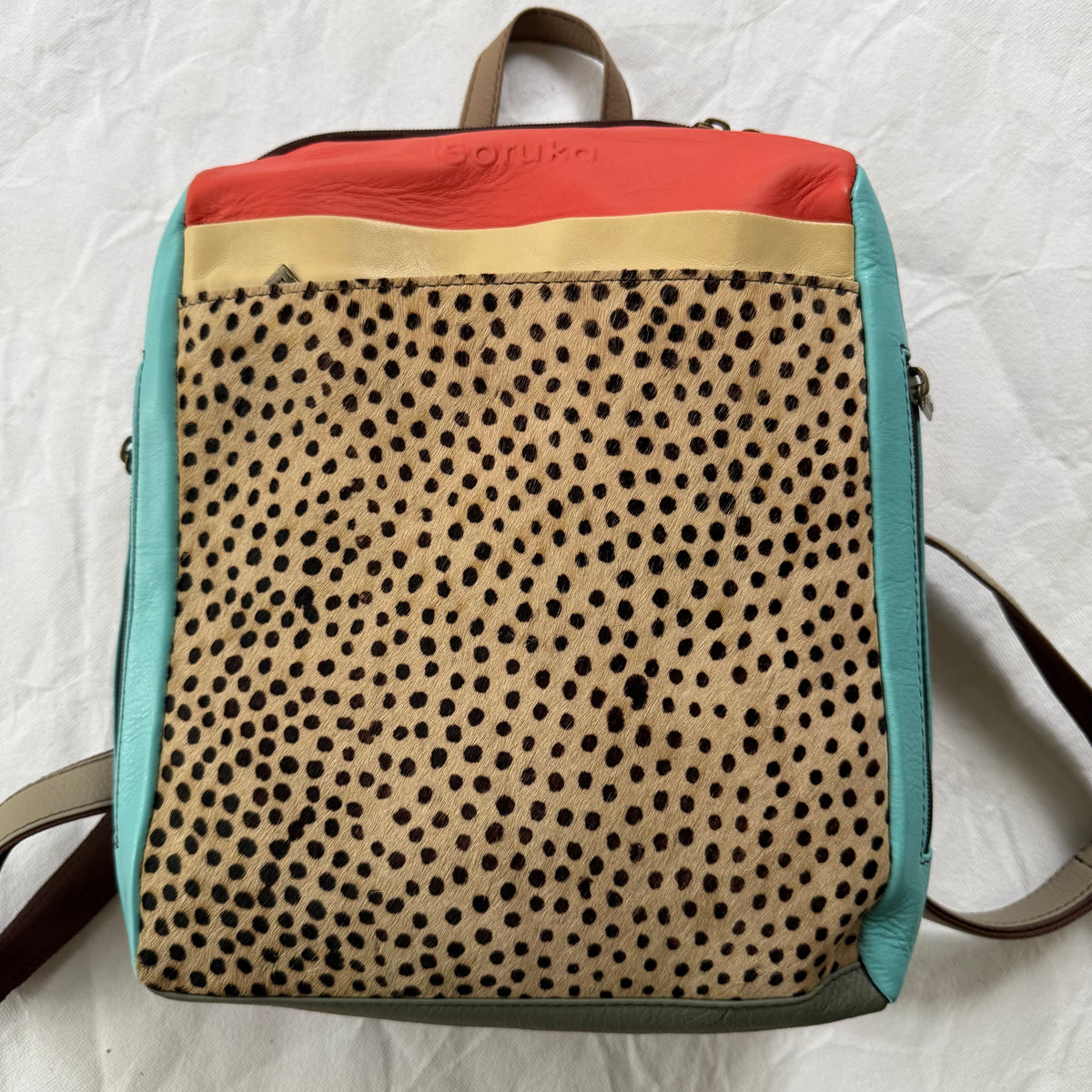 A multicolor leather backpack with a leopard print front panel, a coral top section, a beige stripe, turquoise side panels, and brown leather straps