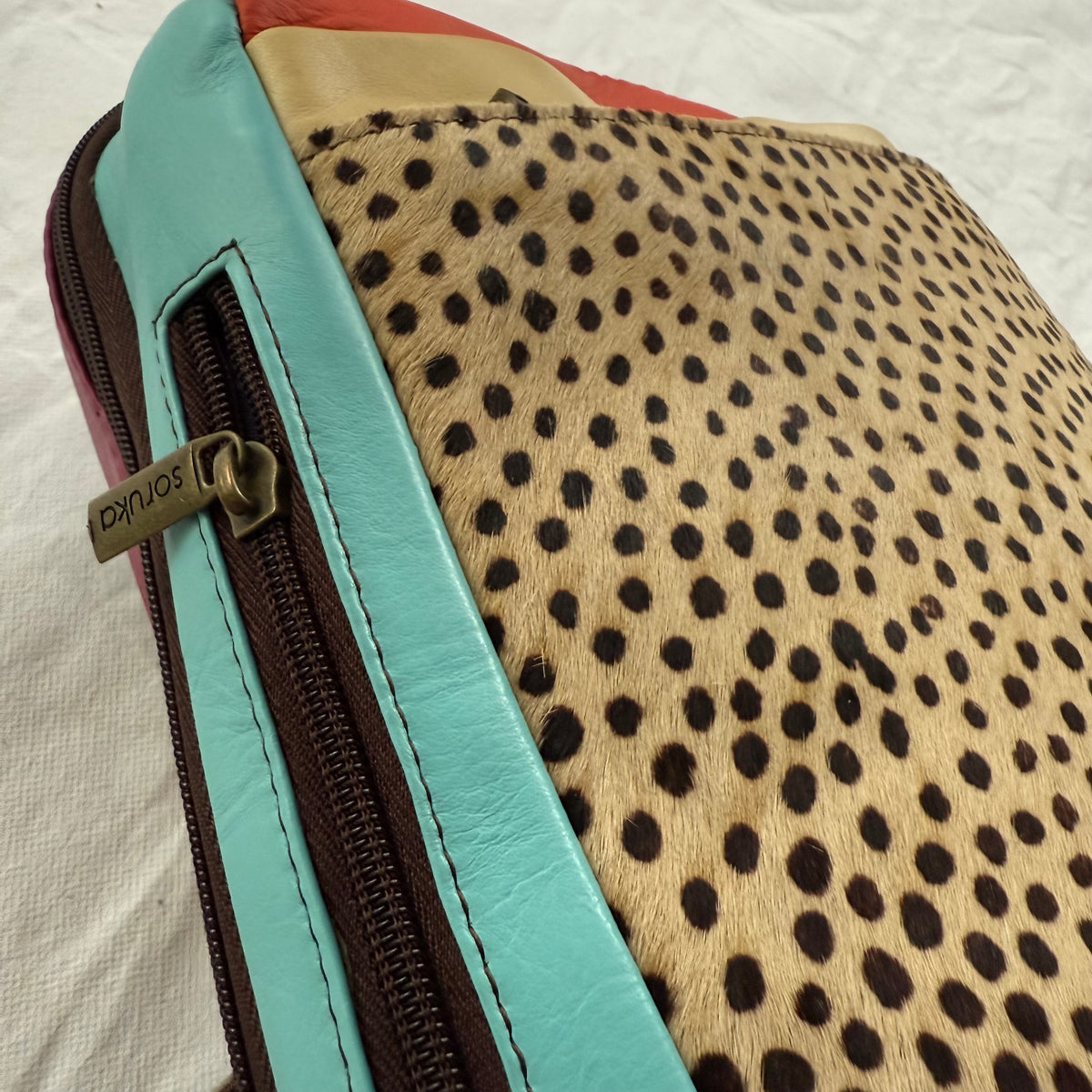 Close-up of side view of backpack featuring a tan and black spotted animal print front panel, a turquoise leather side with and a dark brown zipper with a metal pull.