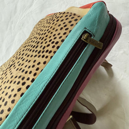 Close-up of side view of backpack showing a leopard print top panel, a turquoise side pocket, a pink base, and a zipper pull with the brand name Soruka.