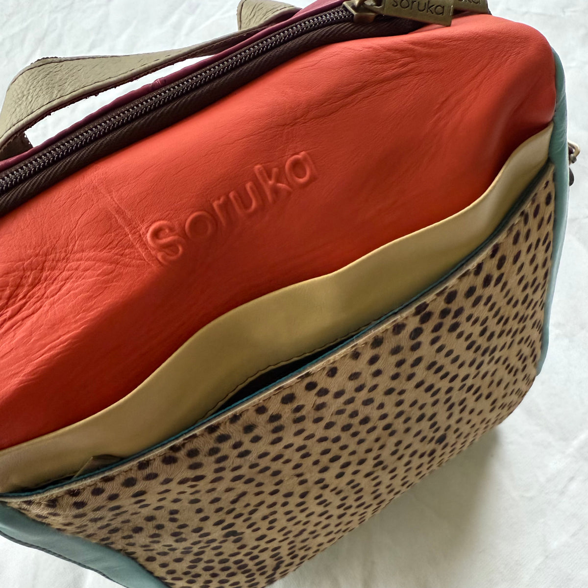 A close-up of a Soruka leather backpack with coral colored top, pale yellow band, and animal print front