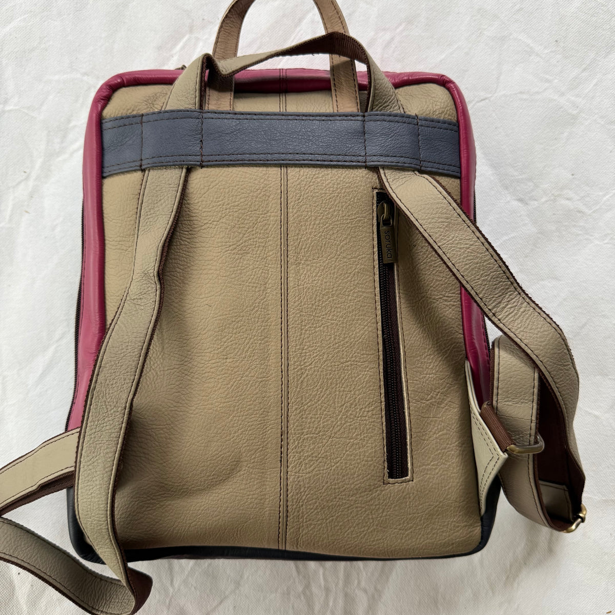 back view of beige leather backpack with a dark gray horizontal strap across the upper back, a pink side panel, and a vertical zippered pocket