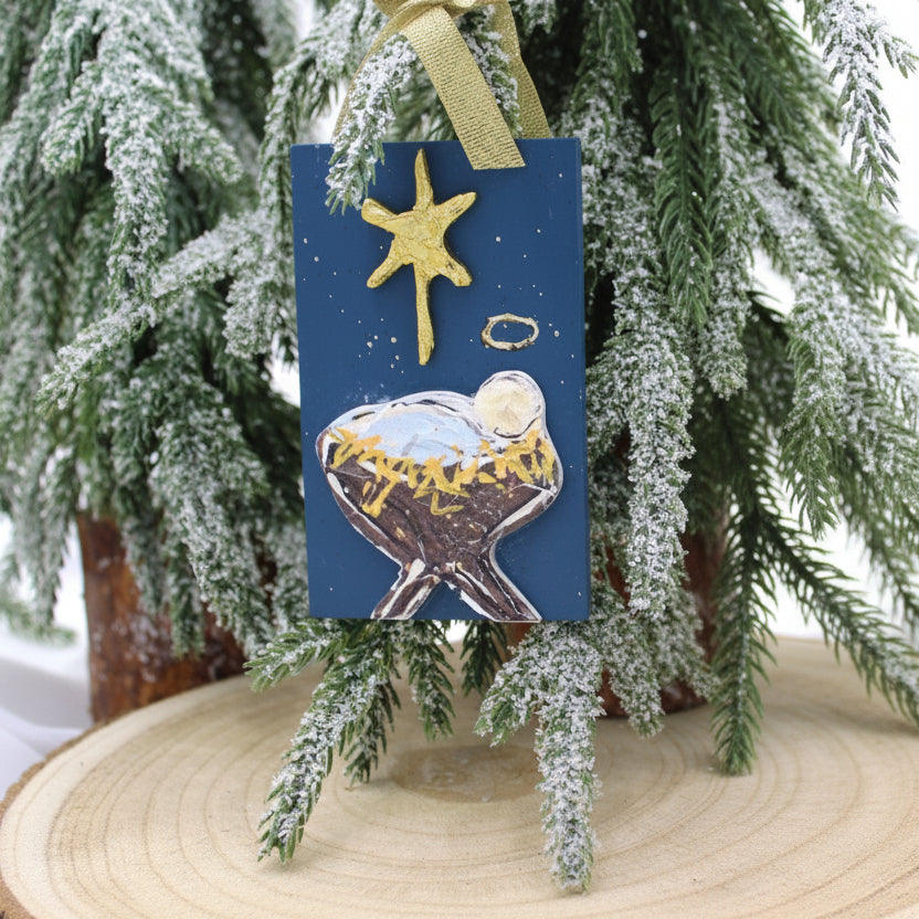 blue ornament of jesus in a manger with a north star above on a flocked tree