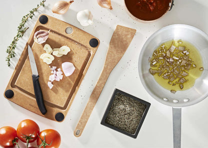 Cooking setup with cutting board, knife, wooden utensil, pan, and ingredients on a white surface