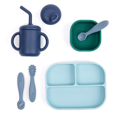 silicone dinnerware for baby, light blue plate, green bowl, blue cup with lids, and 3 utensils arranged on a white background.