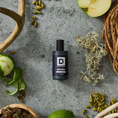 bottle of Buck Moon Proper Cologne on a stone background surrounded by green fruit, seeds, and brown moss