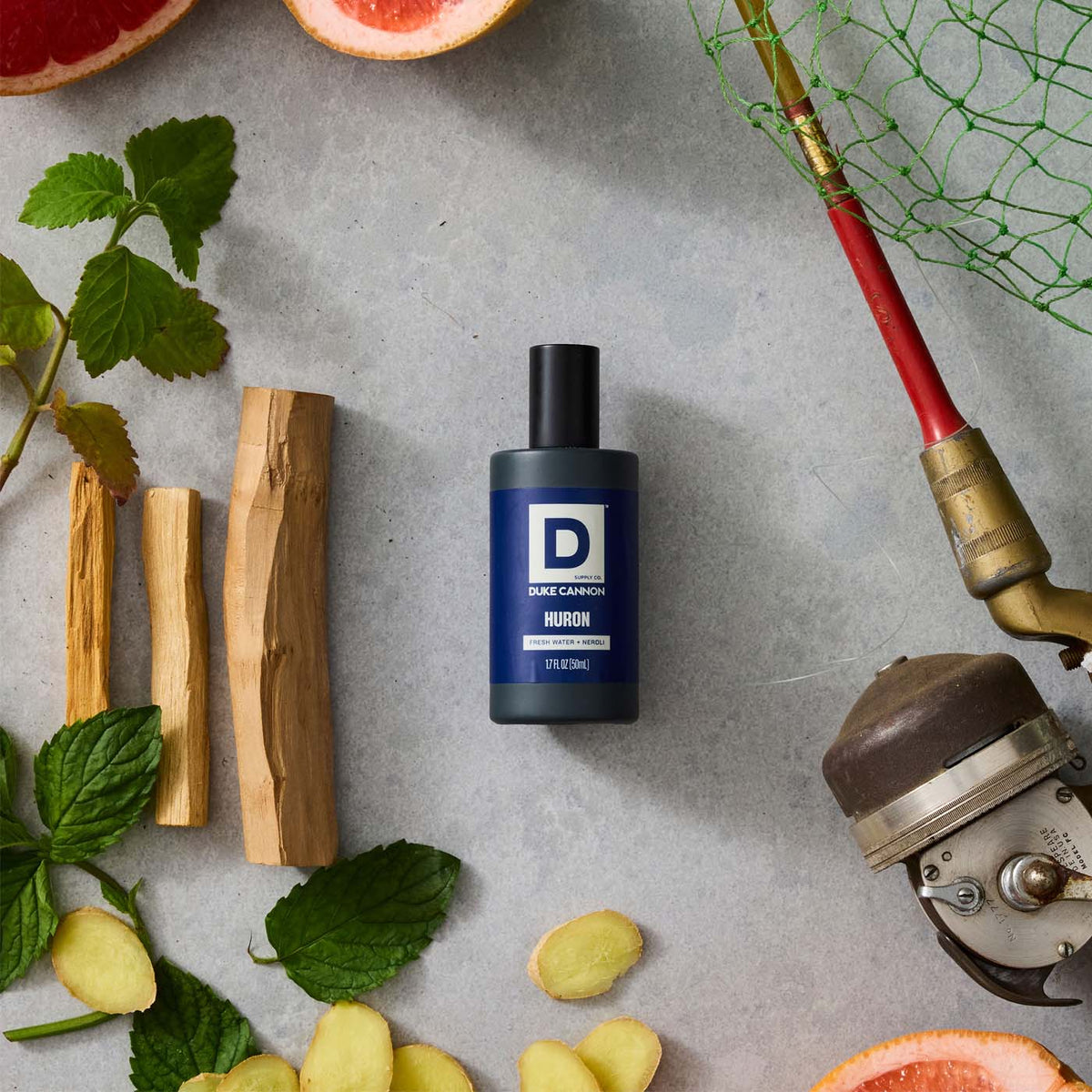 Bottle of Huron Proper Cologne on a stone background surrounded by ginger, citrus, and a fishing rod and net
