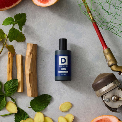 Bottle of Huron Proper Cologne on a stone background surrounded by ginger, citrus, and a fishing rod and net